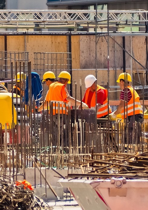 záběr staveniště s pracovníky v oranžových bezpečnostních reflexních vestách a ochranných helmách